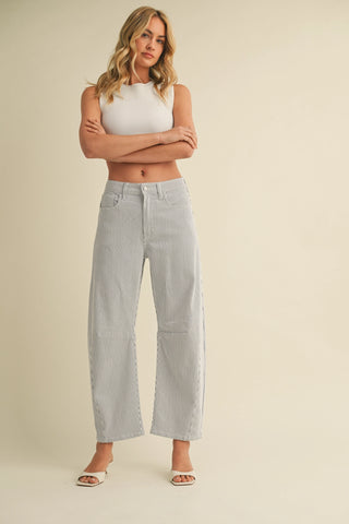 A woman modeling a pair of light gray wide-leg cropped jeans with decorative lace-up detailing on the outer seams, paired with a white sleeveless crop top and white heeled sandals, against a solid beige background.