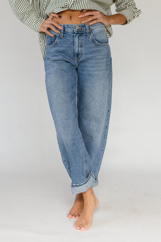 A person wearing light-wash blue jeans with rolled cuffs, paired with a striped shirt, standing barefoot against a plain light gray studio background.