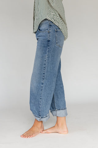 A person shown from the waist down wearing loose, wide-leg blue denim jeans with a cuffed hem, standing barefoot against a plain light gray studio background.