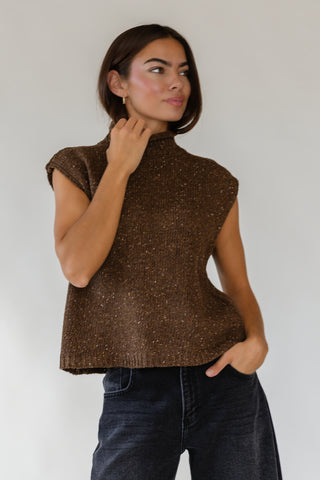 A woman wearing a brown sleeveless knit mock-neck top with subtle flecks, paired with dark blue jeans, standing against a plain light gray background.