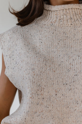 Close-up of a person wearing a sleeveless ribbed knit sweater vest in a warm off-white or cream color with subtle multicolor flecks, featuring a mock neck.