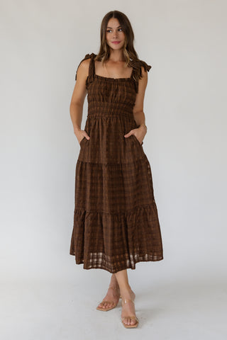 A woman modeling a brown tiered midi dress with a subtle plaid pattern, featuring shoulder tie straps and side pockets, set against a plain light gray studio background.