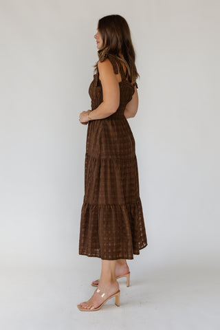 A woman modeling a sleeveless brown tiered maxi dress with a subtle plaid pattern, paired with clear high-heeled sandals, photographed against a plain white studio background.