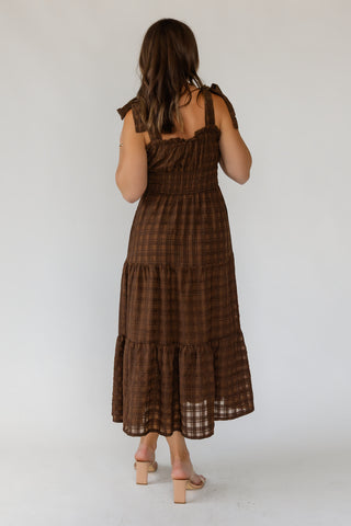 A woman viewed from behind wearing a brown plaid tiered midi dress with tie shoulder straps and beige block-heel sandals, standing against a plain light gray studio background.