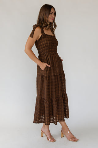 A woman modeling a brown plaid midi dress with tie-shoulder straps and side pockets, paired with clear heeled sandals, against a plain light gray studio background.