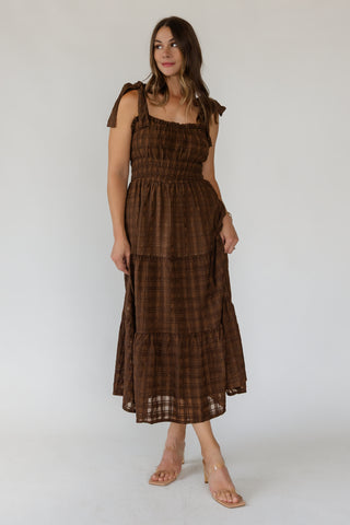 A woman modeling a brown plaid midi dress with shoulder tie details, a smocked bodice, and a tiered skirt, paired with clear heeled sandals against a plain white studio background.