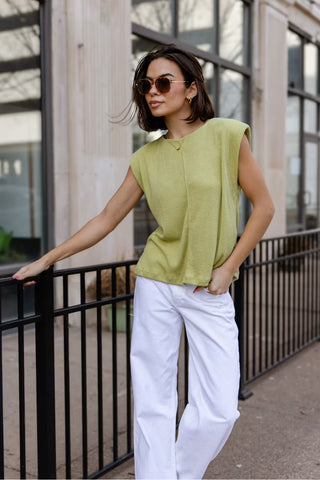Woman wearing a green sleeveless top and white pants standing against a building.
